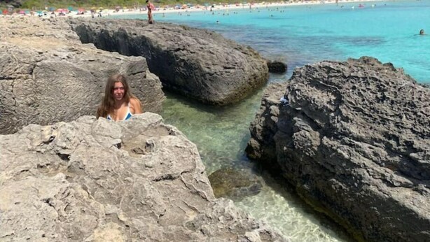 A rocky beach with clear turquoise water and a distant beach area with umbrellas and people.