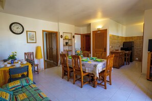 Un coin repas avec une table dressée pour quatre personnes, des chaises, une horloge au mur et une cuisine en arrière-plan.