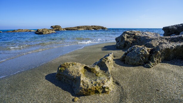 Een strand met helderblauw water, rotsformaties en een kiezelstrand.
