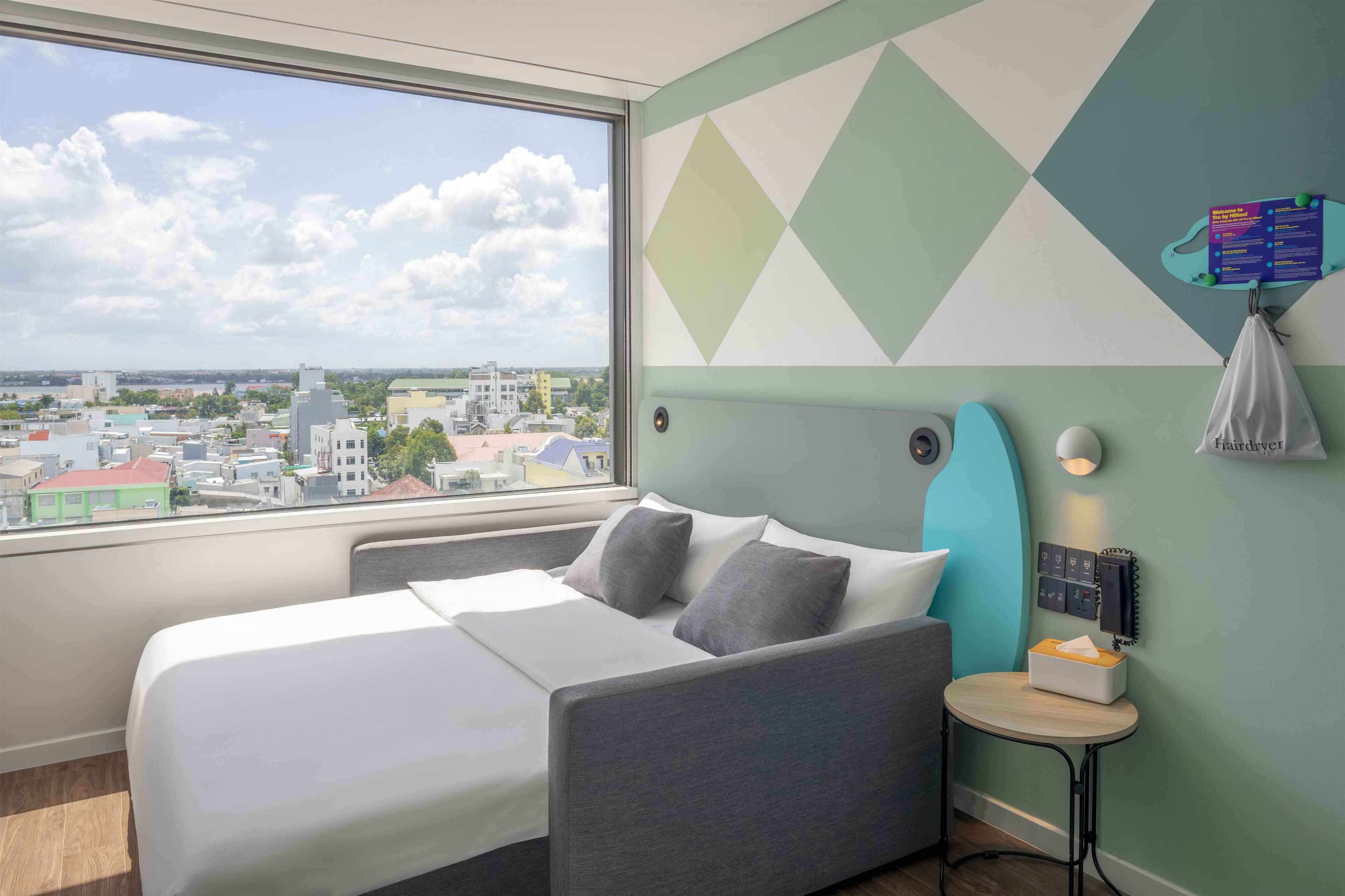 Habitación de hotel con cama, una mesita, luz empotrada en la pared y vista a la ciudad a través de un amplio ventanal.