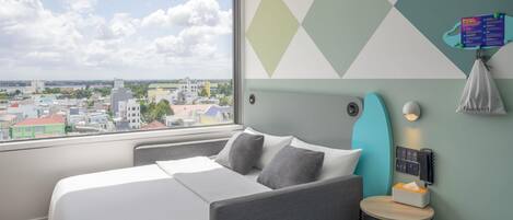 Habitación de hotel con cama, una mesita, luz empotrada en la pared y vista a la ciudad a través de un amplio ventanal.