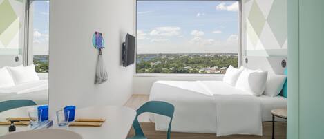 Habitación de hotel moderna con un ventanal que ofrece vistas a la ciudad, una cama con ropa blanca, una mesita con una silla y un televisor montado.