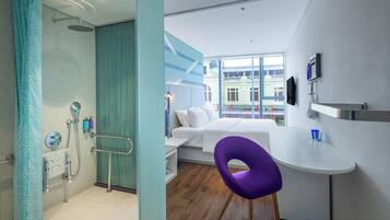 A modern hotel room with a shower, a bed, a small desk, and a view of the city.