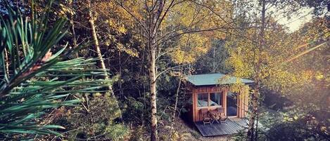 Uma cabana com fachada de madeira, um alpendre com assentos, cercada por uma floresta densa.