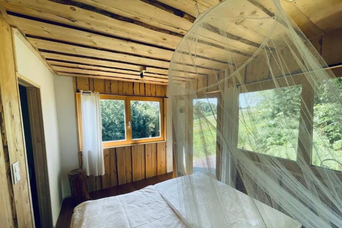 A bedroom with a bed, wooden ceiling, window with curtains, and a view of greenery outside.