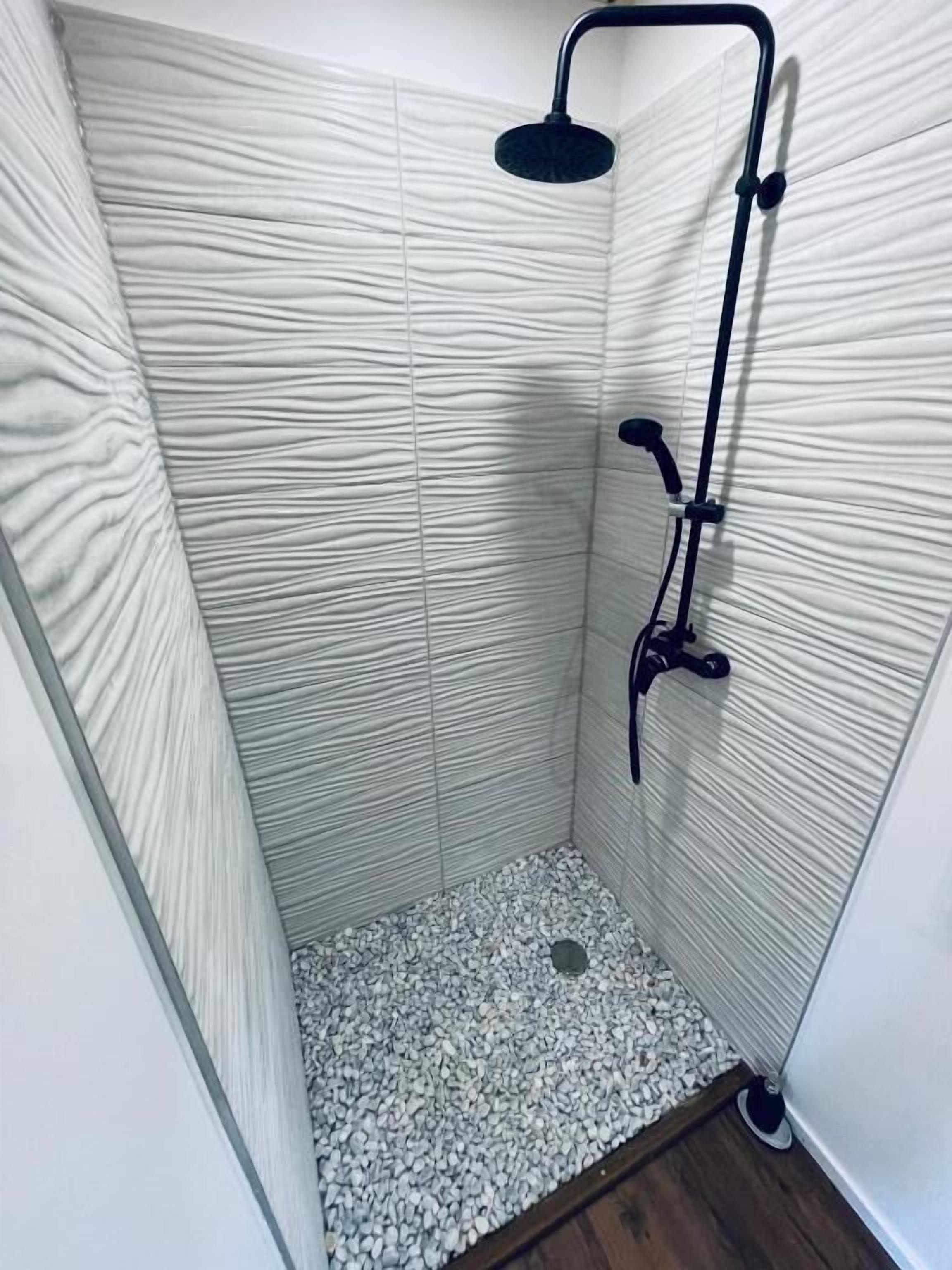 Chuveiro com um chuveiro manual preto, uma torneira fixada na parede e piso de pedras.