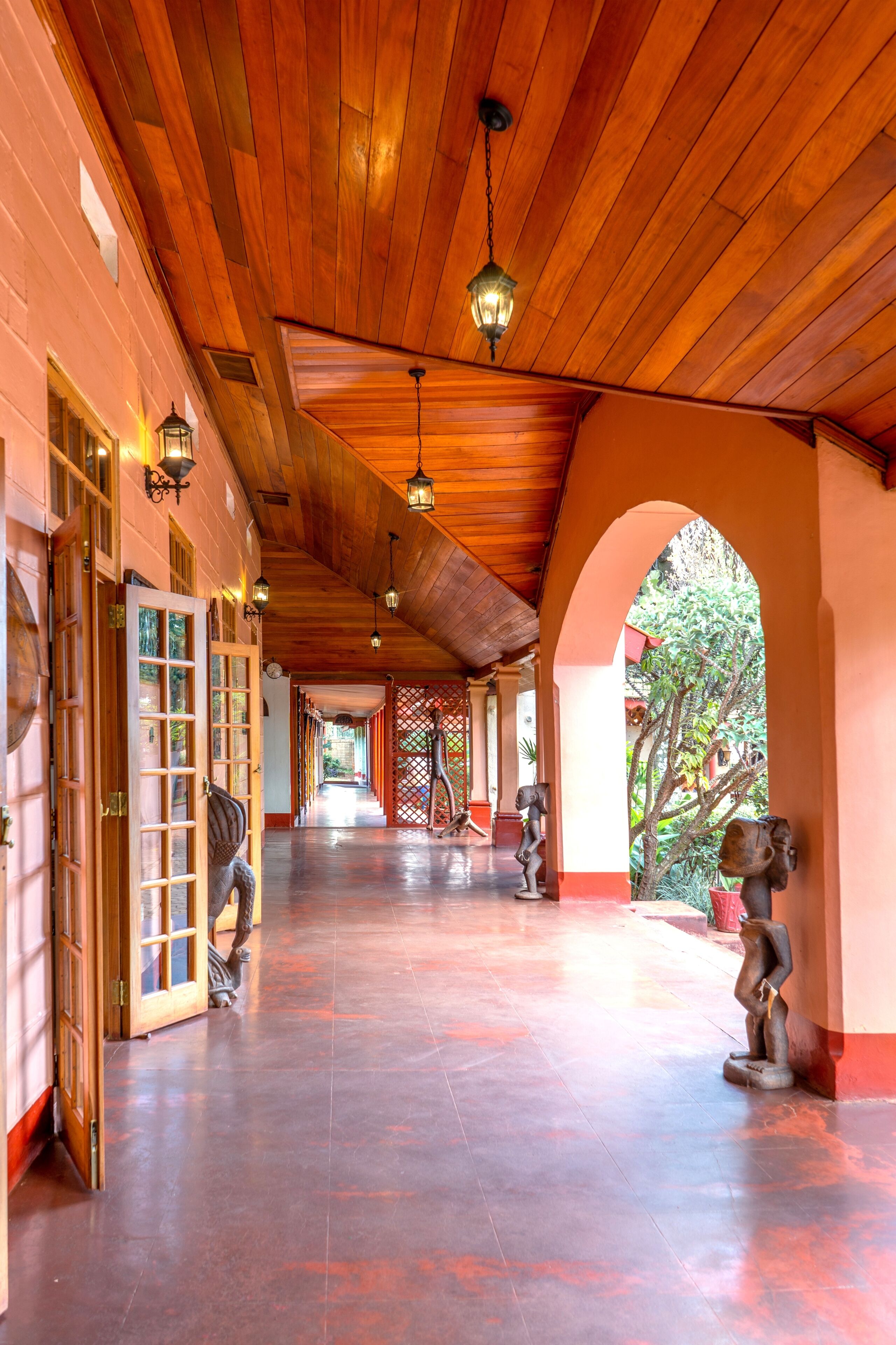 A corridor with wooden ceiling and walls, featuring a series of doors and windows, and a view of the outside through an archway.
