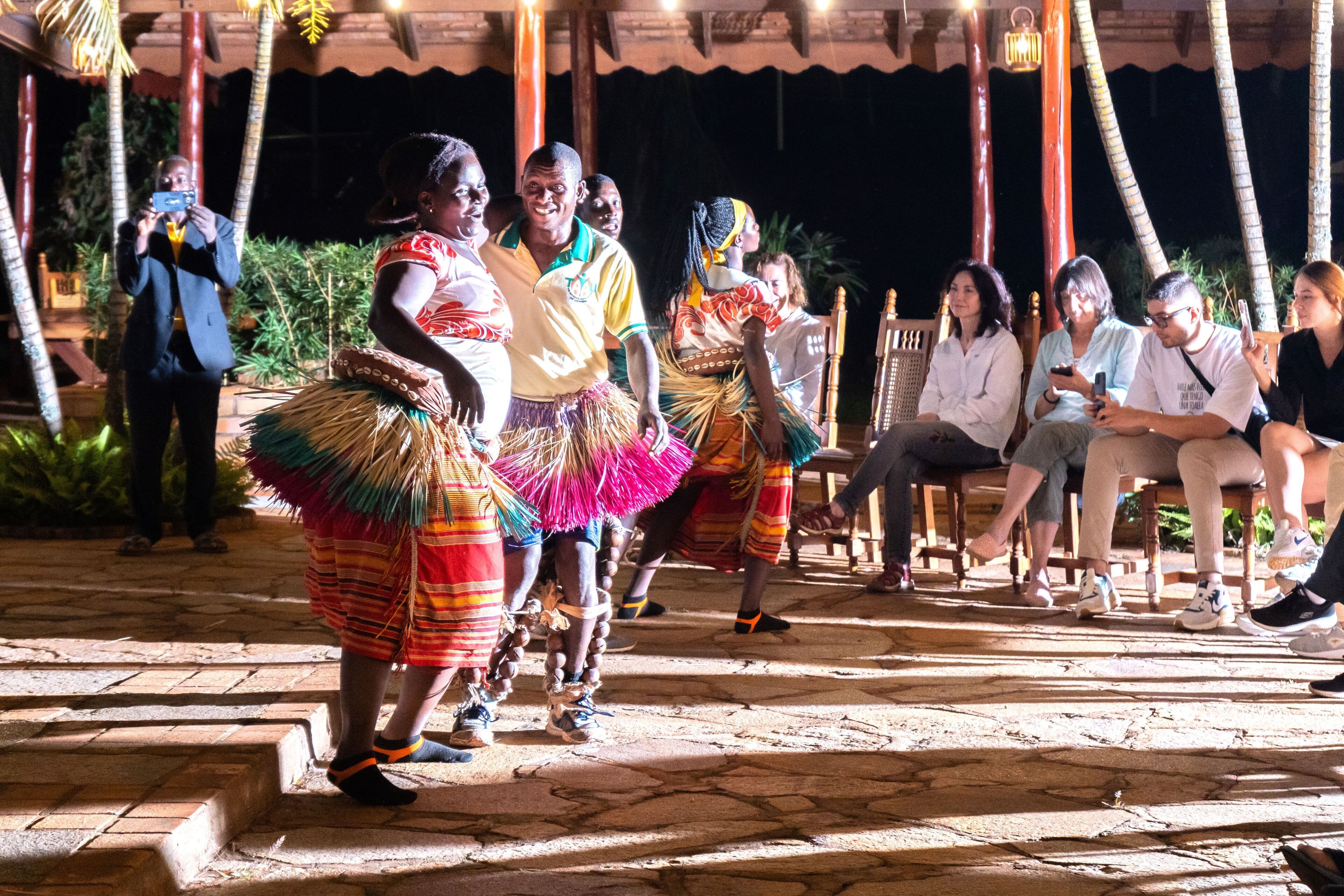 A group of people in traditional attire performing in an outdoor setting with stone flooring and wooden furniture.