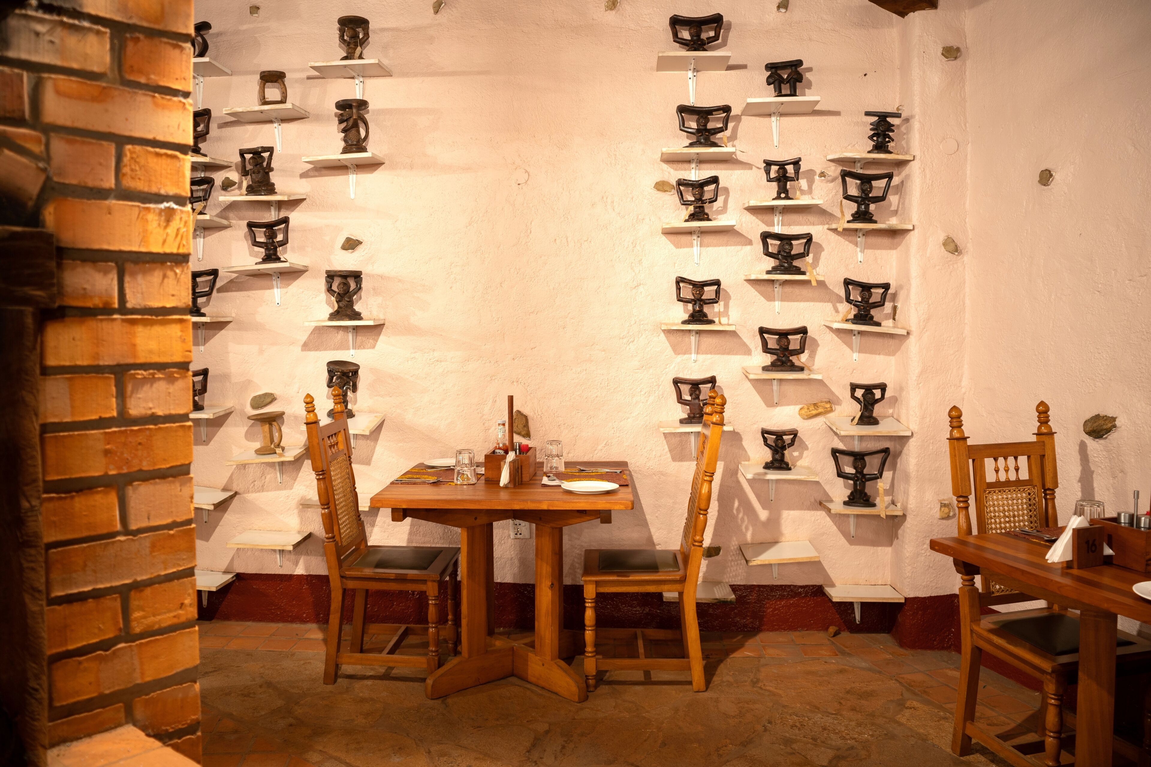 A dining area with a brick wall, wooden table, and chairs, surrounded by shelves with various items.