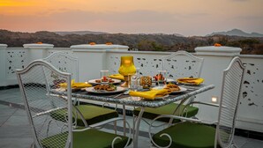 Une table à manger dressée pour deux personnes, avec des chaises à coussins verts, une lampe de table jaune et des assiettes remplies de nourriture. La scène se déroule sur une terrasse d’où l’on aperçoit des collines à l’arrière-plan.
