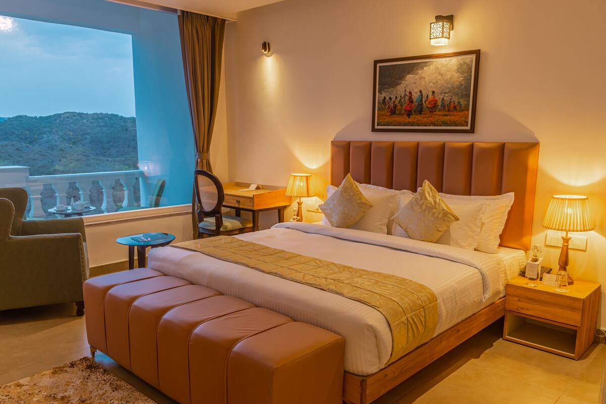 A hotel room with a large bed, a view of the hills, a painting on the wall, and a small table with a lamp.