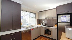 A modern kitchen with dark cabinets, a stainless steel refrigerator, a built-in oven, and a window with blinds.