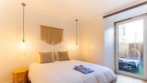 Ein Schlafzimmer mit einem großen Bett, zwei Nachttischlampen, einem Wandbehang aus Makramee und Blick durch eine Glastür auf einen Außenbereich mit Tisch und Stühlen.