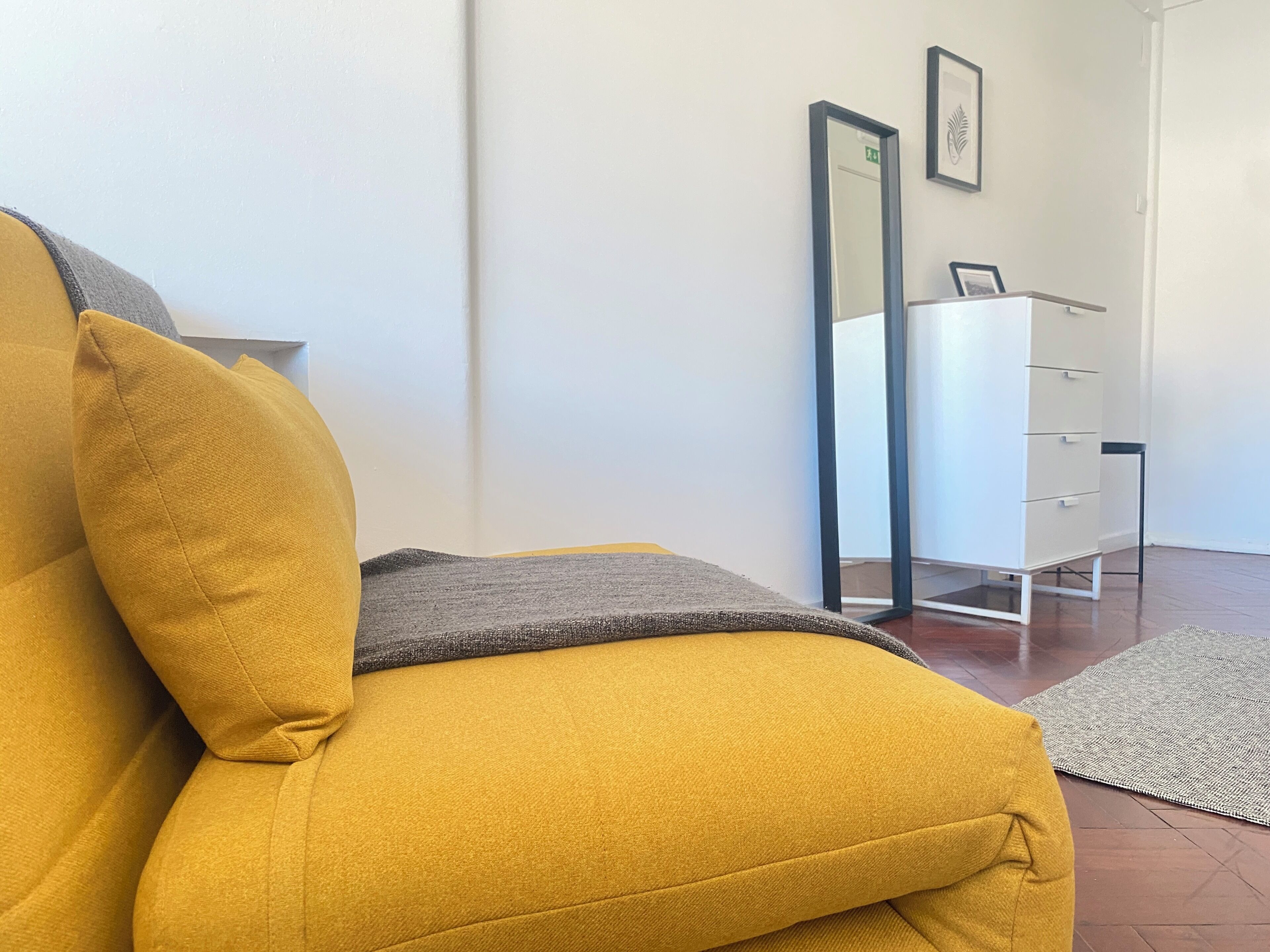 A yellow sofa with a grey throw blanket, a white dresser with drawers, and a mirror on the wall.