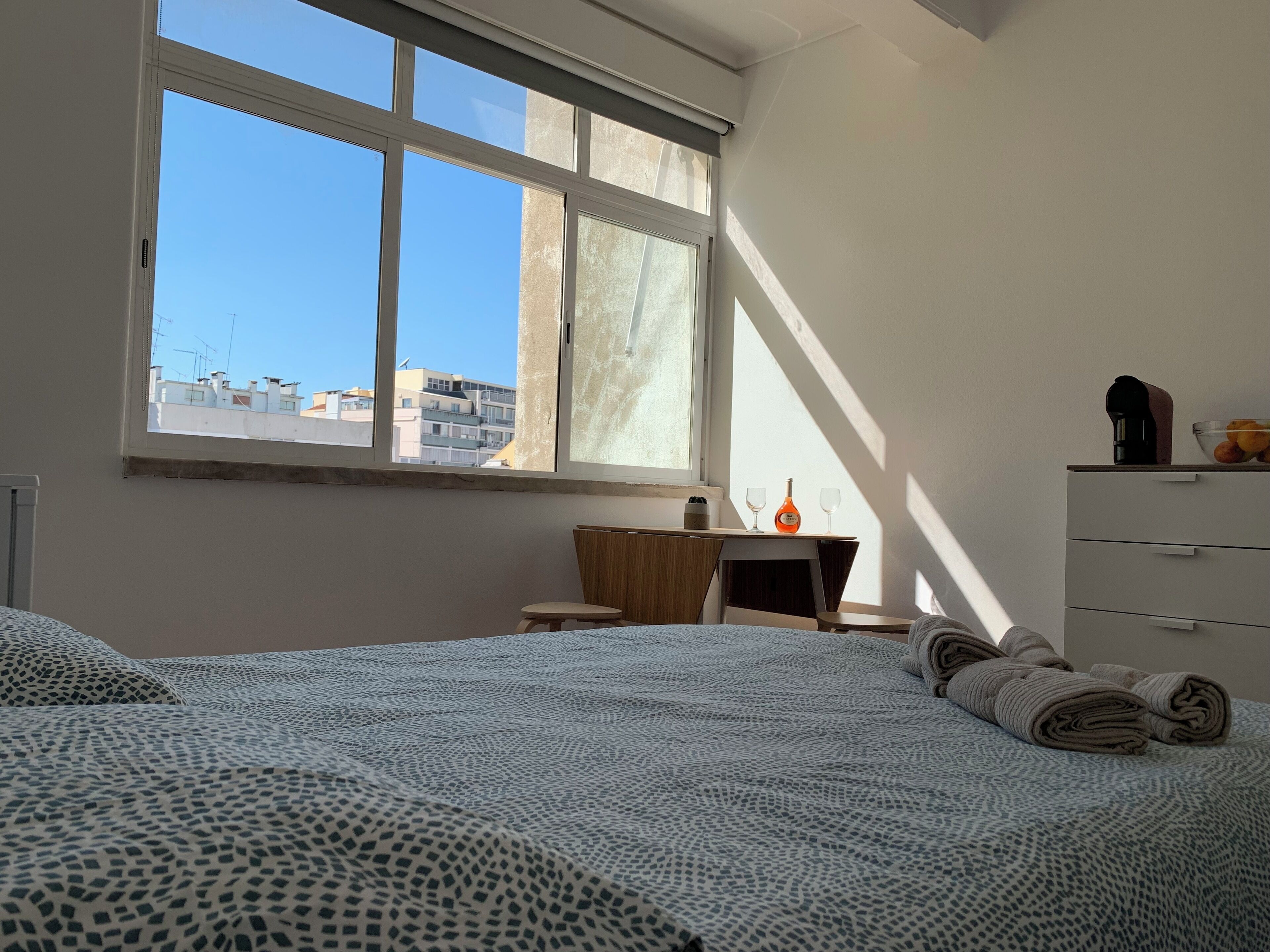 A bedroom with a large window, a desk, a chair, a bed with patterned bedding, and a dresser.