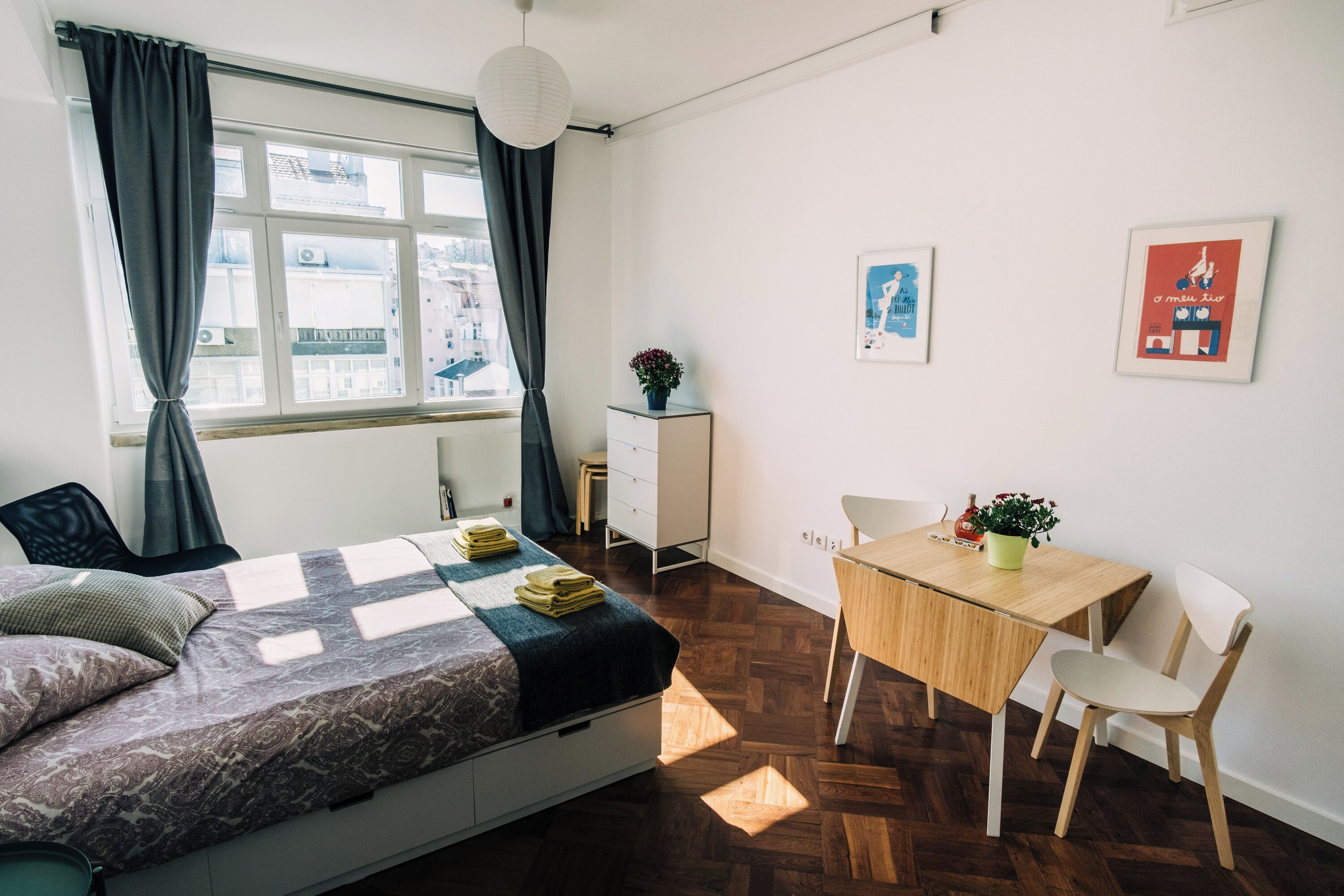 Ein modernes Schlafzimmer mit Bett, Schreibtisch und Stuhl. Ein Fenster mit Vorhängen, eine Topfpflanze und Kunstwerk an der Wand sind zu sehen.