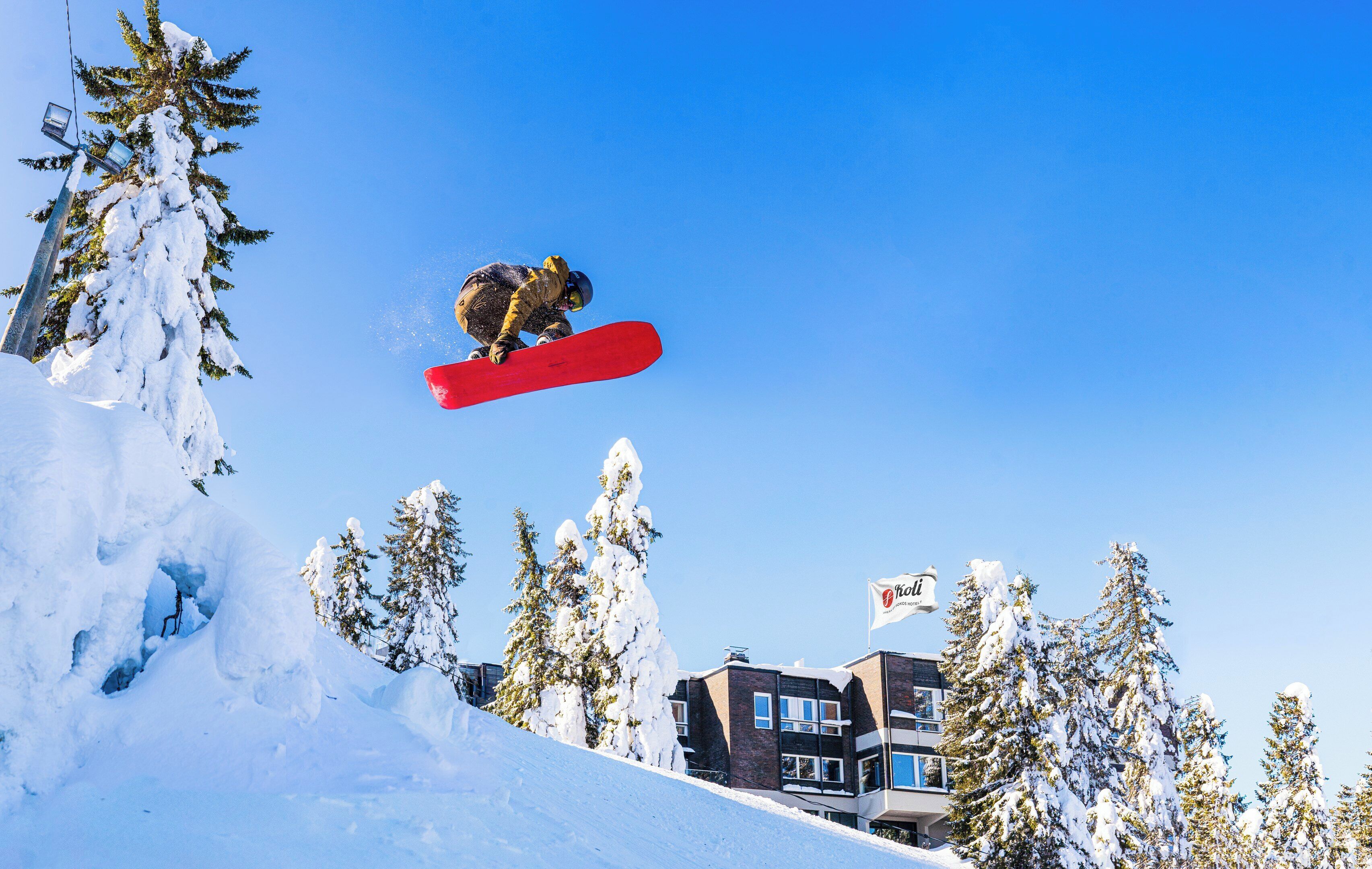 Un snowboarder en pleno salto sobre una ladera nevada, con edificios y árboles al fondo.