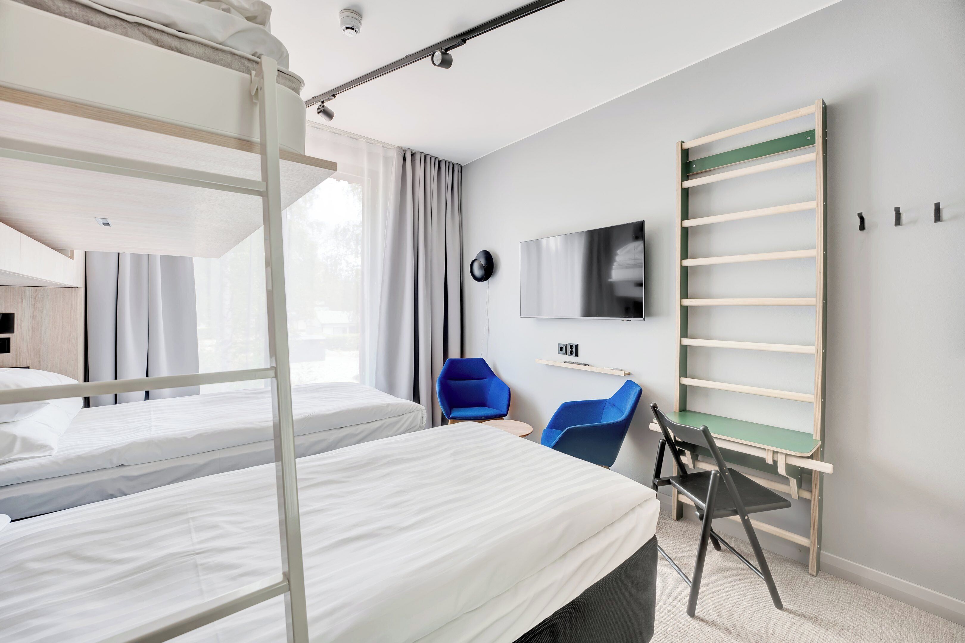 A hotel room with bunk beds, a desk with a chair, a flat-screen TV, and a wall-mounted shelf.