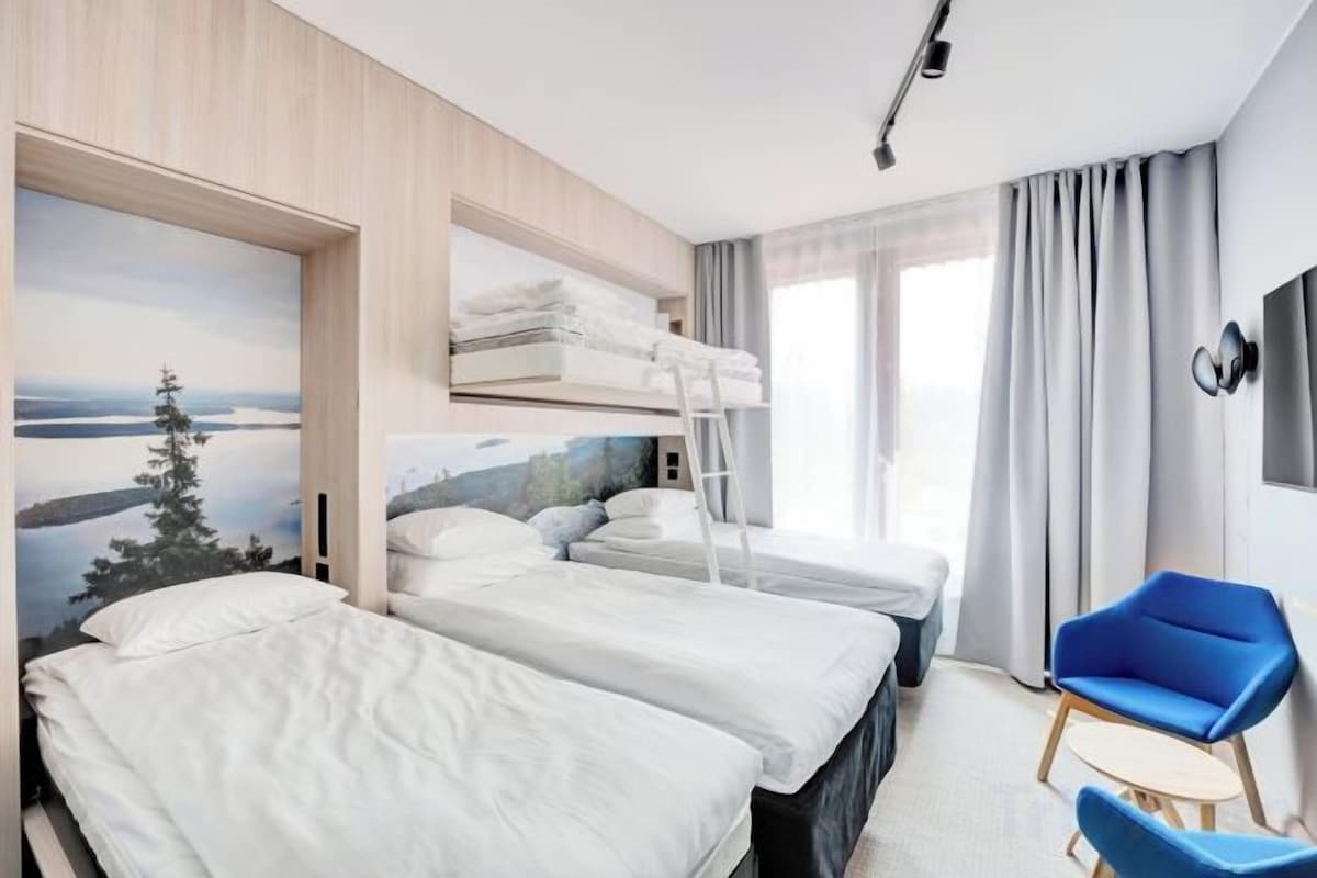 Une chambre d’hôtel avec deux lits, un lit superposé, un bureau, une chaise et une grande fenêtre avec des rideaux.