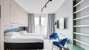 A modern hotel room with a bed, a desk, chairs, a TV, and a large window.