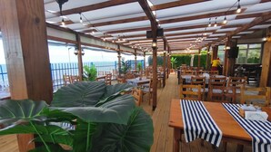 An outdoor dining area with wooden tables and chairs, a view of the sea, and a covered roof structure. - Panorama-apartmentsChantall (Ravda)