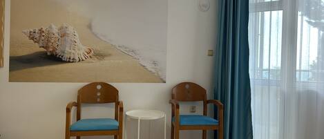 A room with two wooden chairs with blue cushions, a small white table, a bed with a beige quilt, and a large painting of a seashell on the wall.