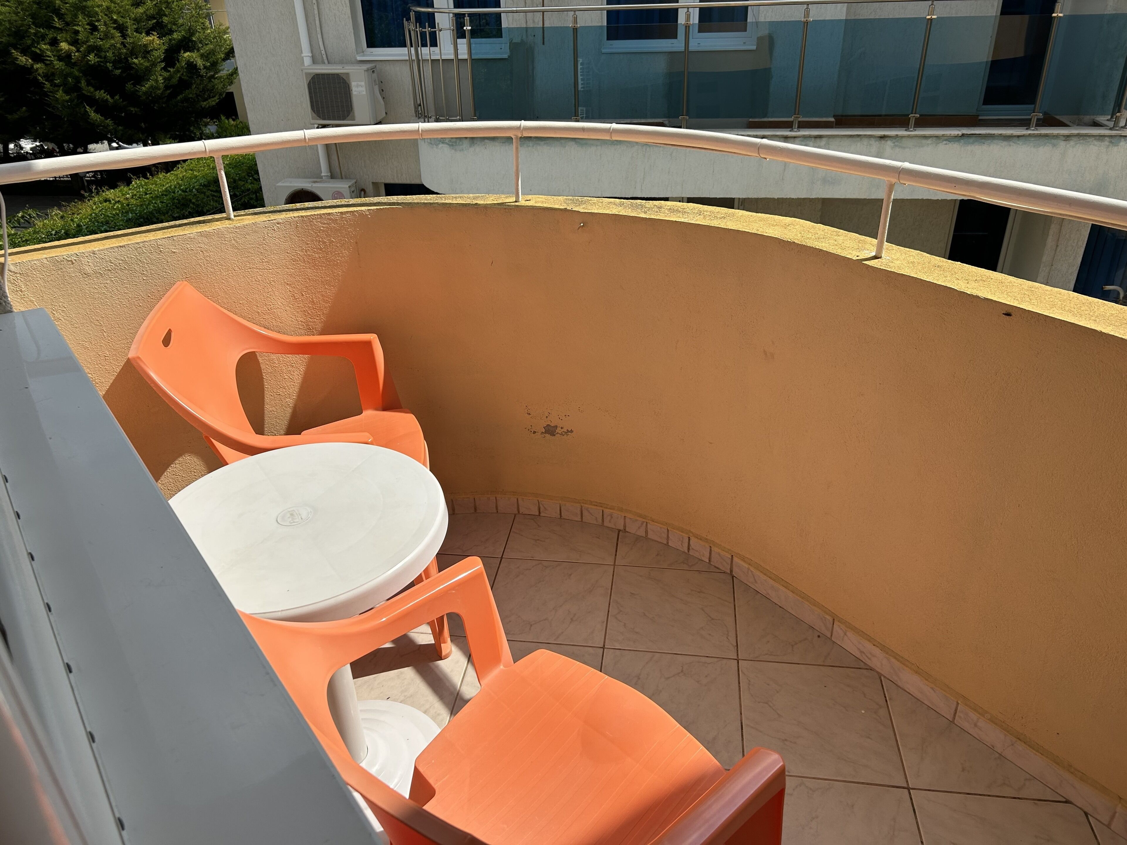 A curved outdoor seating area with orange chairs and a white table, surrounded by a yellow wall and a metal railing.