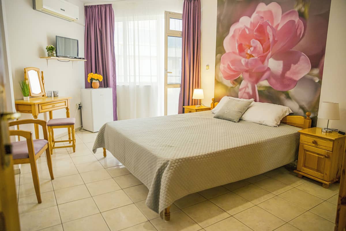 A hotel room with a bed, a desk with a chair, a television, and a large floral painting on the wall.