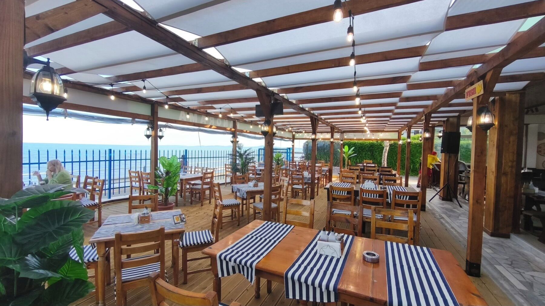 Un restaurant en plein air avec des tables et des chaises en bois, une vue sur la mer et un espace couvert doté d’un plafond en bois.