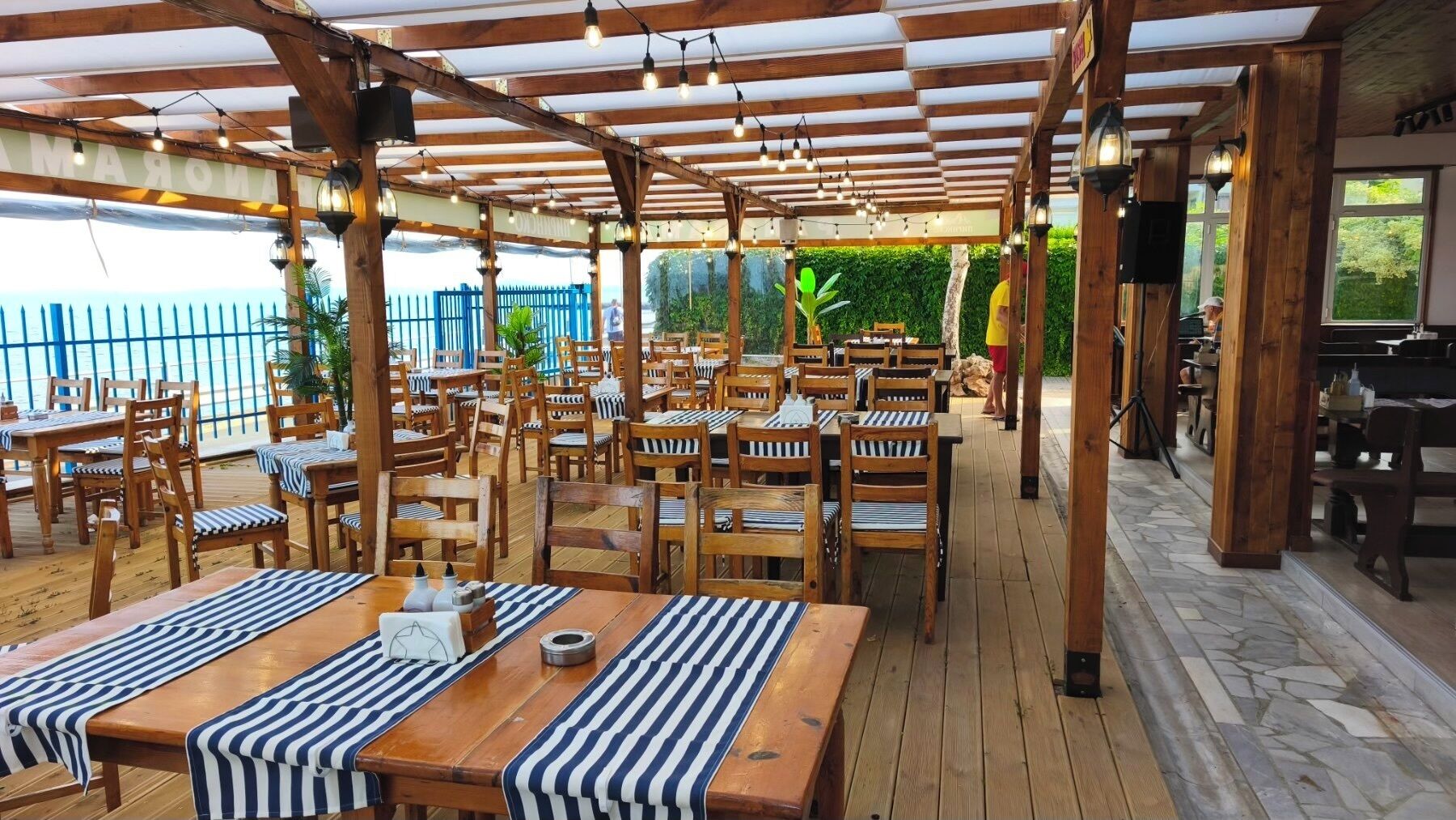 An outdoor dining area with wooden tables and chairs, covered by a wooden pergola with string lights.