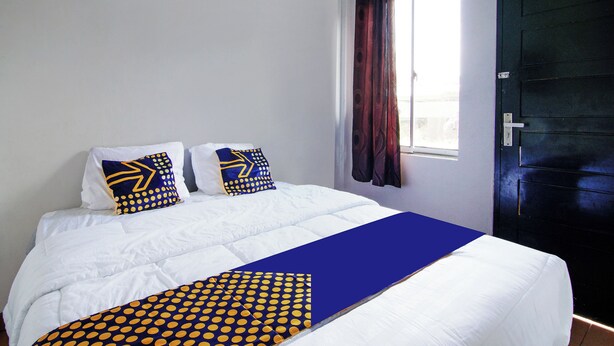 A neatly made bed with white bedding and blue and yellow polka dot pillows, a dark door, and a window with curtains. - Hotel O Oklahoma Homestay Syariah (Pekanbaru)
