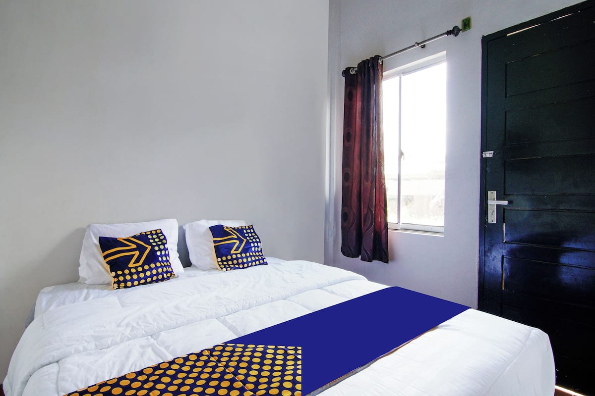 Ein ordentlich gemachtes Bett mit weißer Bettwäsche und Kissen mit blauen und gelben Punkten, eine dunkle Tür und ein Fenster mit Vorhängen.