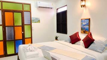 A hotel room with a bed, a laptop, a wall-mounted air conditioner, a door with colorful glass panels, and a painting on the wall.