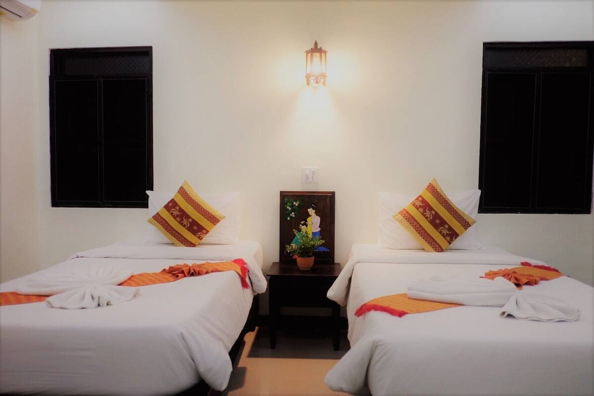Ein Hotelzimmer mit zwei Betten, jeweils mit weißer Bettdecke und einem Zierkissen. Dazwischen befindet sich ein kleiner Tisch mit einem gerahmten Bild.