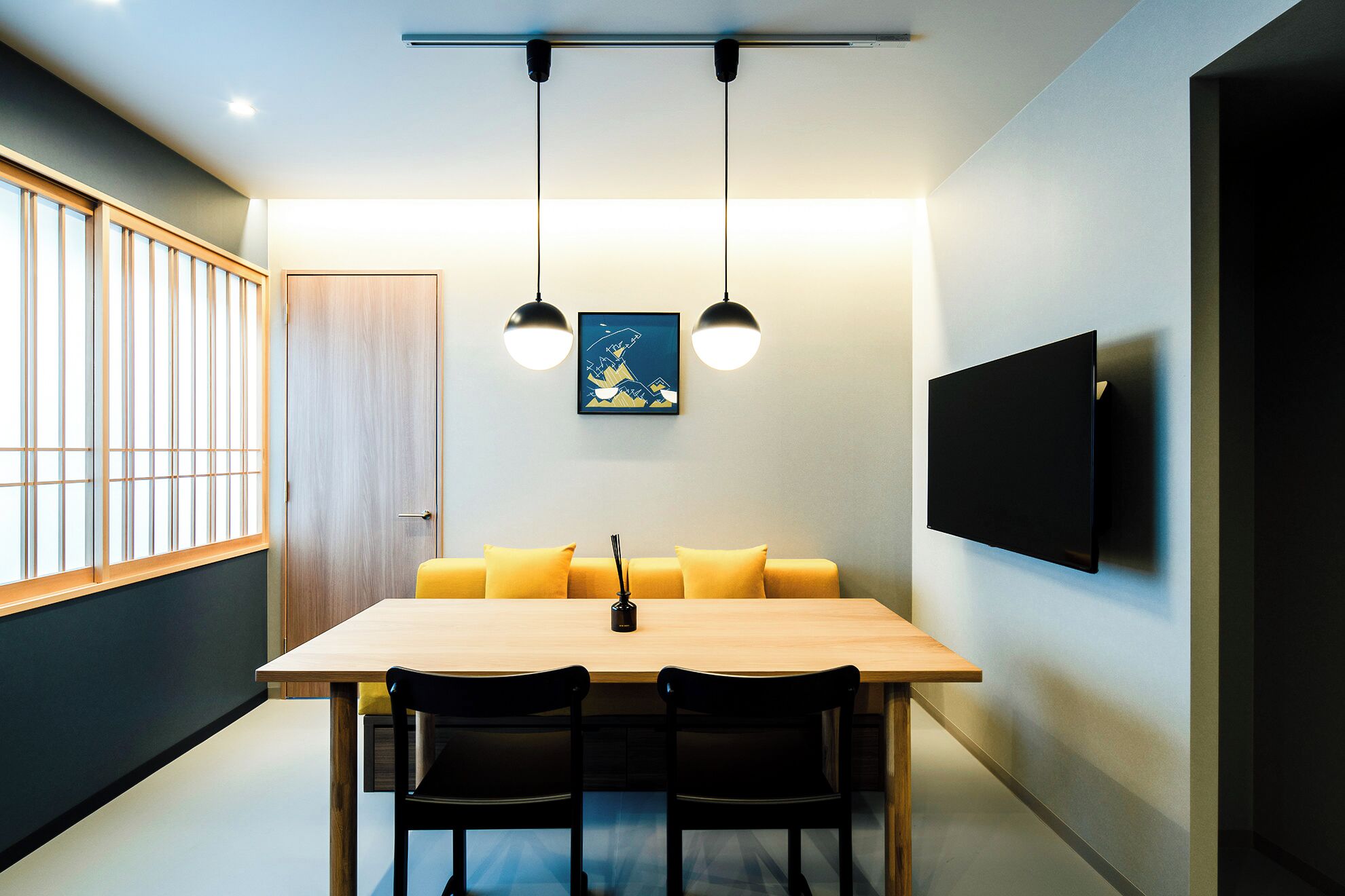 Un comedor moderno con una mesa de madera, dos sillas negras, un sofá amarillo, una lámpara negra y un televisor montado en la pared.