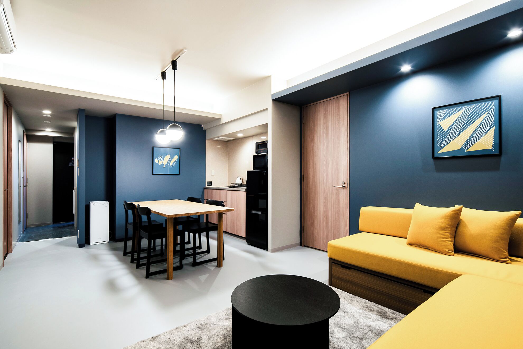 A modern living room with a sofa, a dining table with chairs, a kitchen area with appliances, and a hallway.