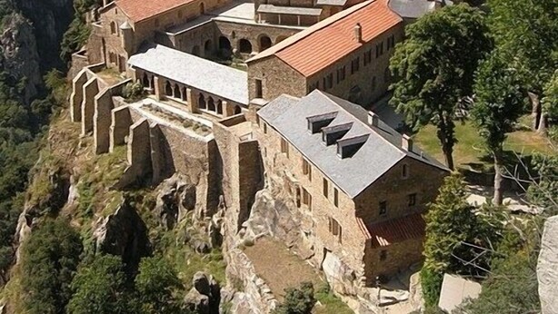A historic stone building with multiple levels and a tower, surrounded by lush greenery and mountains.