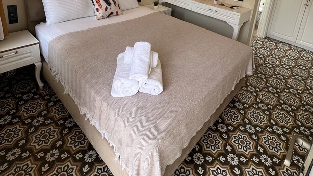 A neatly arranged hotel room with a large bed, a bedside table, a dresser, a mirror, and a patterned floor. - Disa Hotel (Cesme)