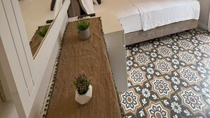 A hotel room with a bed, a bedside table with a plant, a mirror, a TV mounted on the wall, and patterned flooring. - Disa Hotel (Cesme)
