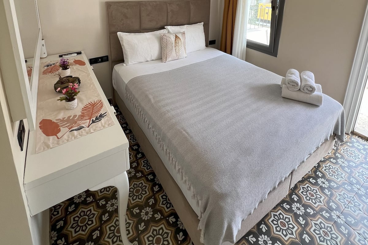 Une chambre d’hôtel avec un lit, un bureau, un miroir, une fenêtre avec des rideaux et un sol à motifs.
