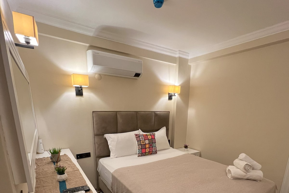 Une chambre d’hôtel avec un lit, des tables de chevet, un bureau, une lampe, une plante et des carreaux de sol à motifs.