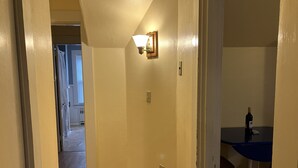 A hallway with a wooden floor, a light fixture on the wall, and a table with a bottle and a glass. - 2-Bedroom Apt Sleeps 7! - 1/4 mile to Beach! (Hampton)