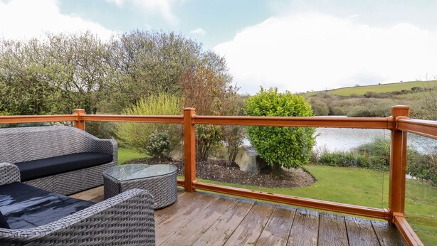 Um deck de madeira com móveis de vime e uma mesa de vidro, com vista para um lago e vegetação.