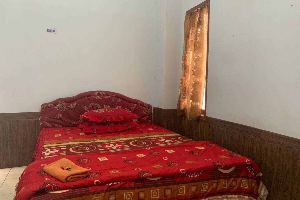 A single bed with a red patterned bedspread, a wooden headboard, and a red pillow.