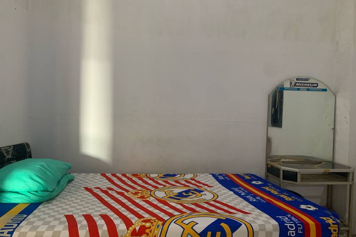 A bed with a checkered bedspread featuring sports team logos, a teal pillow, a small bedside table, and a mirror on the wall.