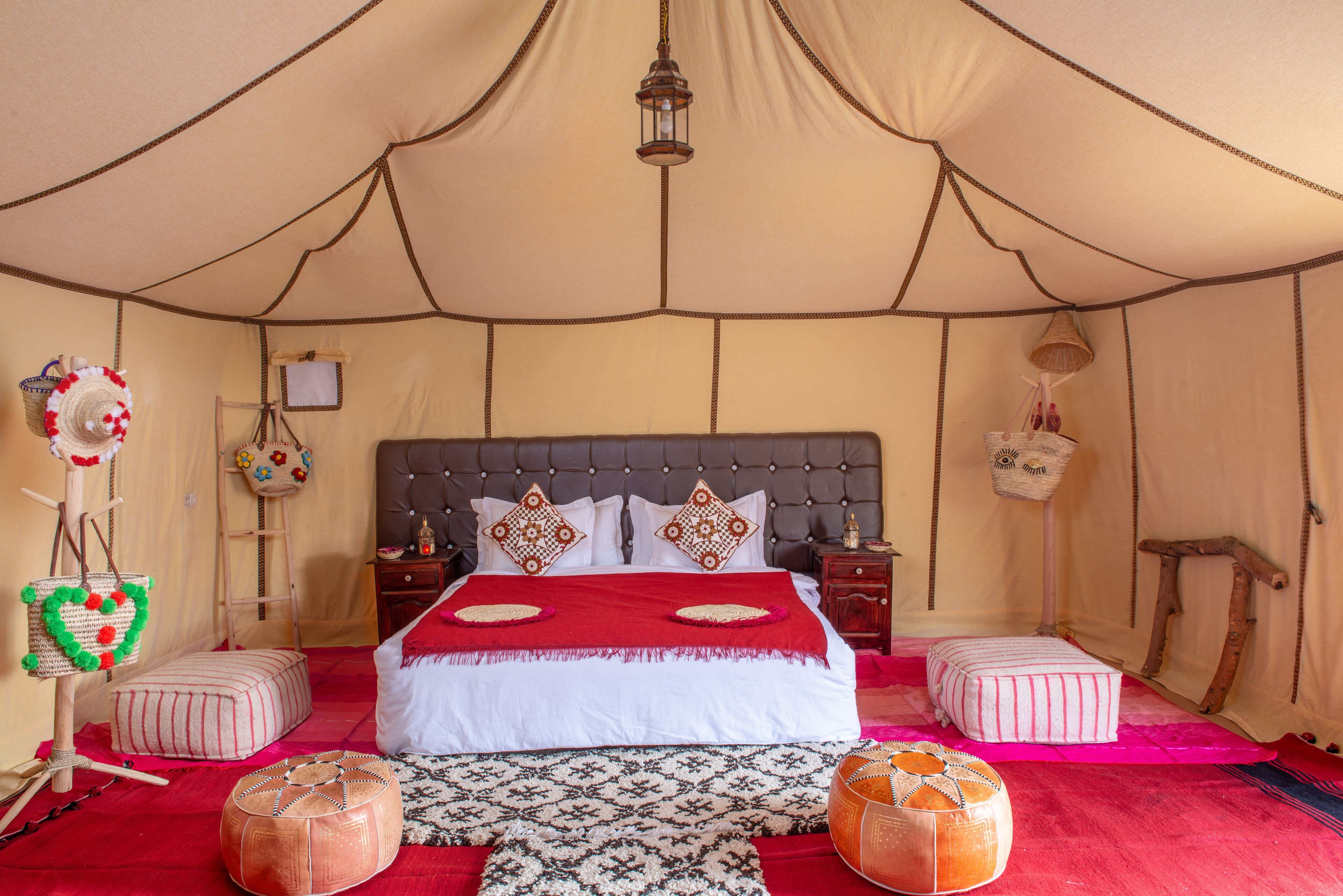 A tented room with a large bed, two ottomans, a hanging lantern, and a small table with a lamp.