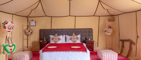 A tented room with a large bed, two ottomans, a hanging lantern, and a small table with a lamp.