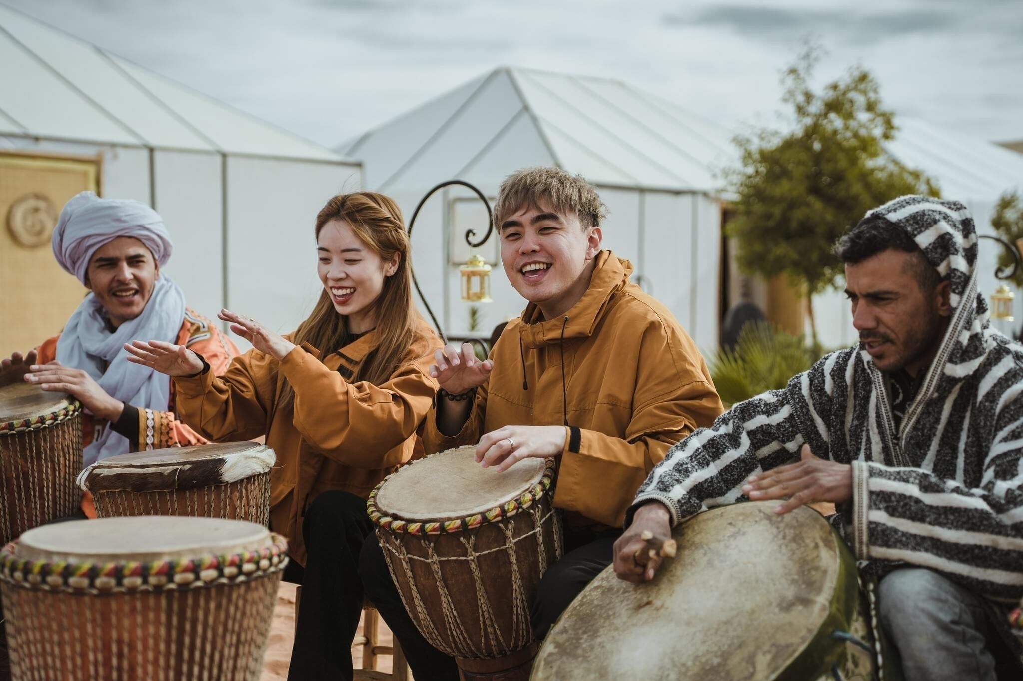 A group of people playing drums in an outdoor setting with a white tent in the background.