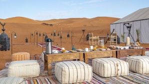 A desert camp with a dining table set for breakfast, surrounded by ottomans and a tent.