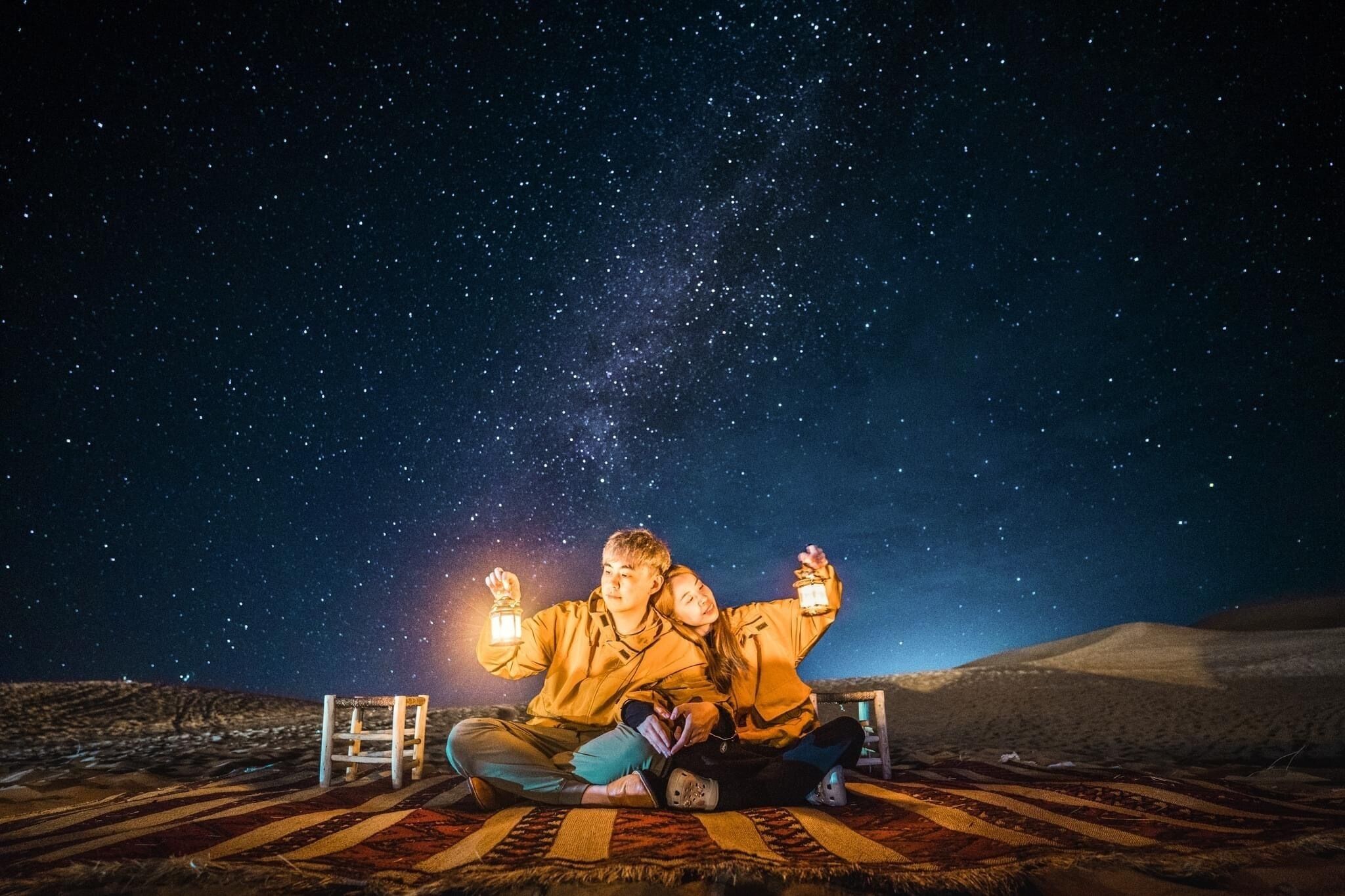 Dos personas sentadas sobre una alfombra estampada bajo un cielo estrellado, sosteniendo faroles.
